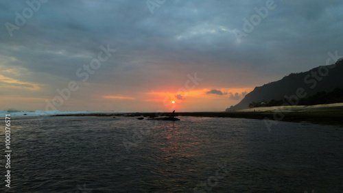 A man sits on a rock in the sea at sunset, admiring the vibrant Bali sky. The waves gently crash around him, creating a moment of calm and reflection.