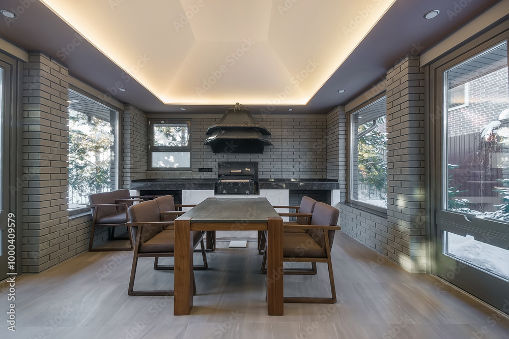 Fototapeta premium The interior of the insulated terrace with an installed barbecue oven and an exclusive hood design. Outside the window is a winter landscape in the rays of the setting sun.