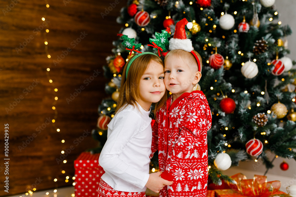 Young boy and girl in crimson and white nightwear with playful holiday headbands are standing beside the Christmas tree. Young boy and girl in crimson and white nightwear with playful holiday headbands are standing beside the Christmas tree.