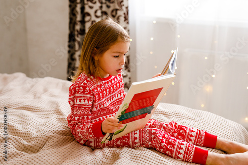 A little girl wearing holiday sleepwear is on the bed, reading a big book