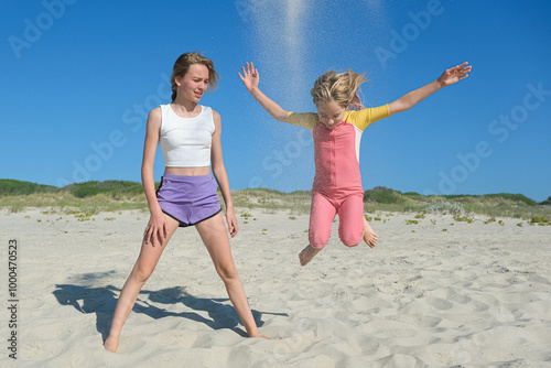 Two girls are playing on a sandy beach. One is jumping joyfully as sand flies up, while the other stands nearby, smiling. The clear blue sky and greenery create a peaceful summer backdrop.