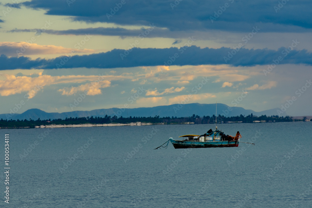 A rustic fishing boat anchored on calm waters with a scenic backdrop of distant mountains under a cloudy sky at sunset. A rustic fishing boat anchored on calm waters with a scenic backdrop of distant mountains under a cloudy sky at sunset.