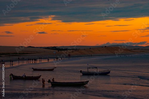 Vibrant sunset over a beach with boats and people silhouetted against orange and blue hues, capturing the lively essence of Jericoacoara. photo