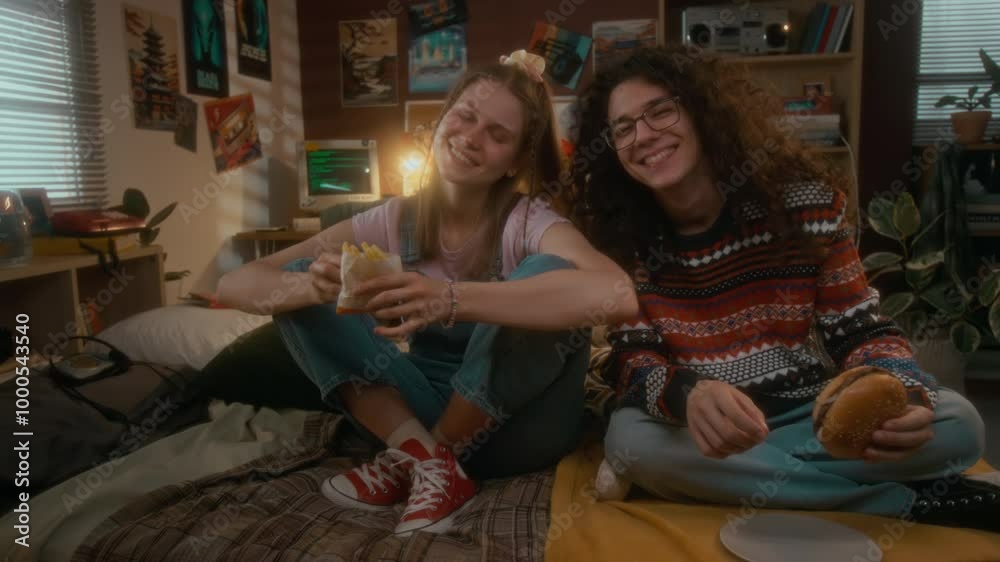 Full handheld portrait of cheerful gen Y Caucasian couple posing with burger and french fries in bed in retro-styled bedroom, looking at camera, nostalgic and carefree 90s Full handheld portrait of cheerful gen Y Caucasian couple posing with burger and french fries in bed in retro-styled bedroom, looking at camera, nostalgic and carefree 90s
