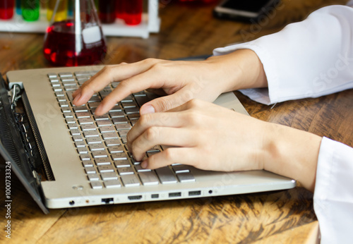 Close up male lab assistant chemist using laptop for data entry and in other hand holding test tube with red blood sample while sitting in central laboratory.