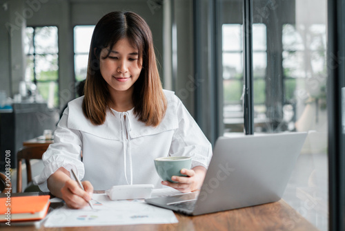 A young woman working remotely from home, using a laptop and holding a coffee cup, in a modern and bright home office environment.