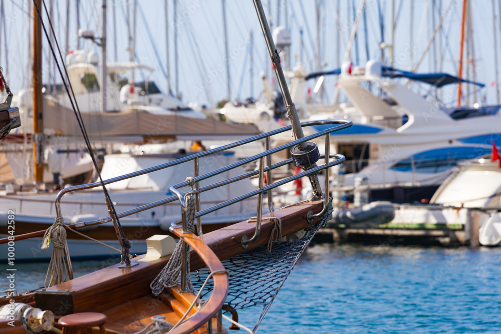 Fototapeta premium Bow of wooden sailboat in marina, surrounded by luxury yachts. Polished wood and chrome details, clear blue water and yachts in blurred background