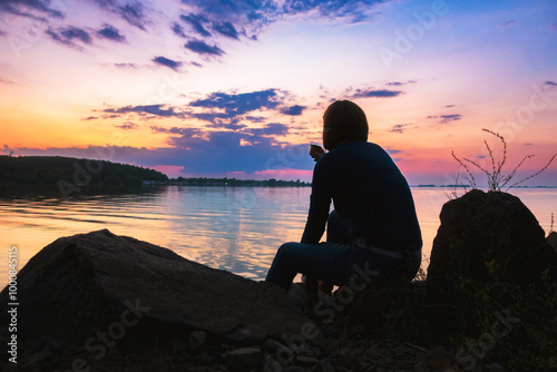 The woman enjoys the sunset by the river sitting on the stones with a cup of tea.