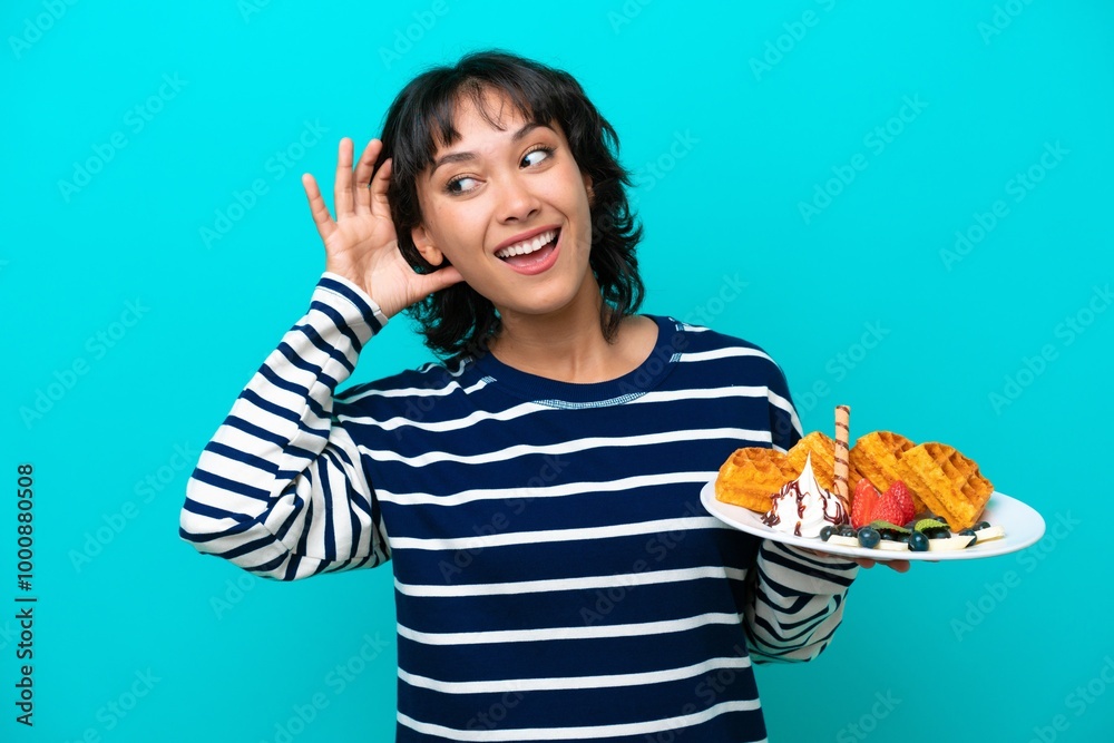 Young Argentinian woman holding waffles isolated on blue background listening to something by putting hand on the ear Young Argentinian woman holding waffles isolated on blue background listening to something by putting hand on the ear
