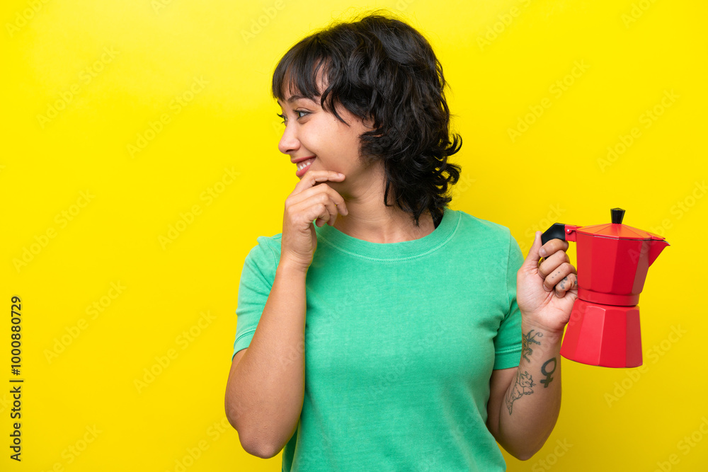 Young Argentinian woman holding coffee pot isolated on yellow background thinking an idea and looking side Young Argentinian woman holding coffee pot isolated on yellow background thinking an idea and looking side