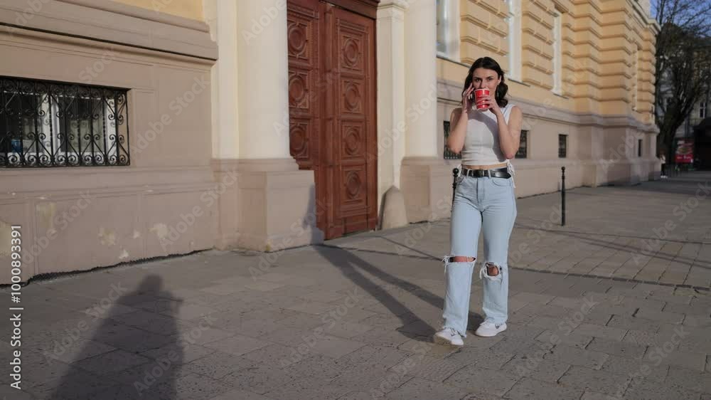 Dialing mobile, Coffee stroll, Young woman. Strolling through city, young woman in jeans and white top engaged in phone conversation, carrying red cup of coffee. Dialing mobile, Coffee stroll, Young woman. Strolling through city, young woman in jeans and white top engaged in phone conversation, carrying red cup of coffee.
