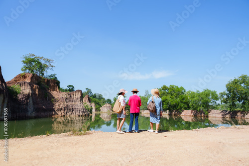 Three elderly Asian women standing near a calm lake, dressed in casual outfits and wide-brim hats. The natural scenery of cliffs and water create a peaceful and serene outdoor setting. photo