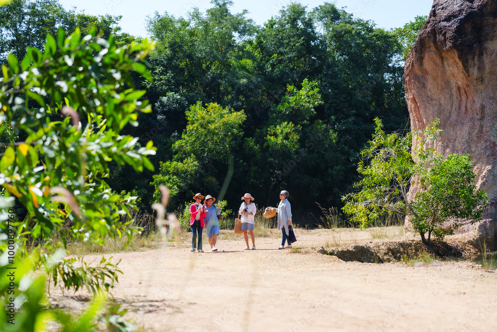 Fototapeta premium Four elderly Asian women happily walking and enjoying each other's company in a scenic outdoor environment. Casual summer outfits and wide-brim hats reflect their relaxed, joyful nature exploration.