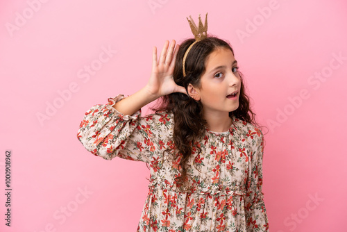 Little caucasian princess with crown isolated on pink background listening to something by putting hand on the ear