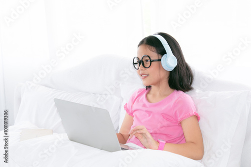 Adorable Asian girl concentrate online education class on laptop, kids lie down on bed watching game or social media connect to friends, smart school girl study e-learning using headphone technology