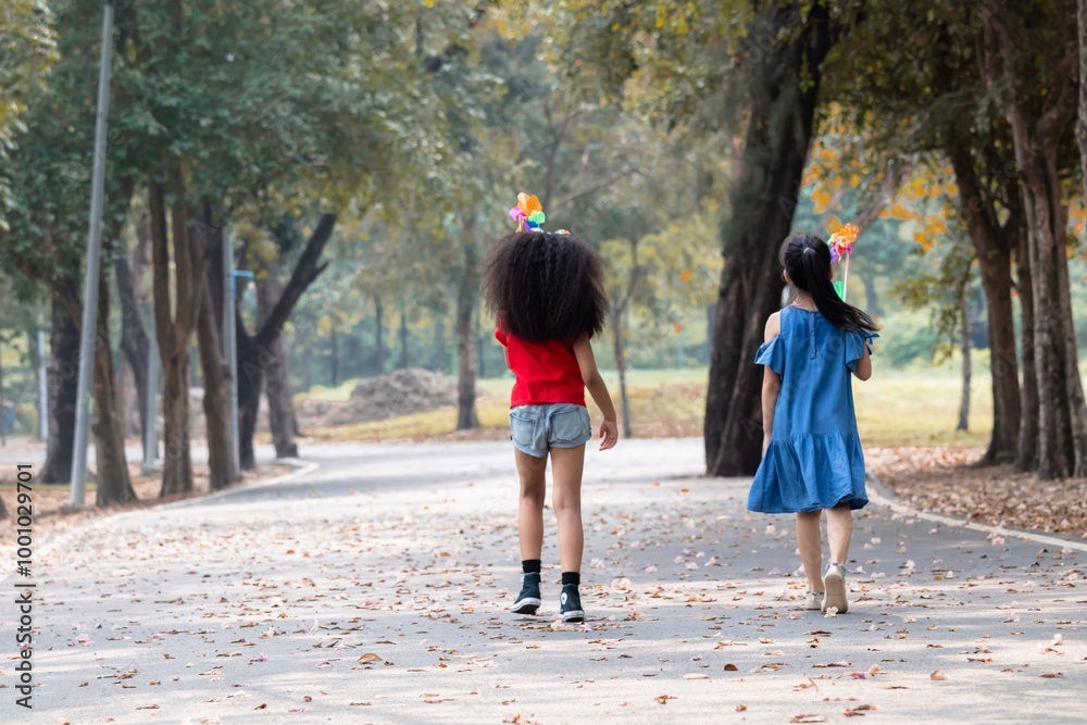 Two multicultural school girls, African-American and Asian kids, walking together hand-in-hand in the park, The moment captures their strong friendship spent weekend free time in natural outdoor Two multicultural school girls, African-American and Asian kids, walking together hand-in-hand in the park, The moment captures their strong friendship spent weekend free time in natural outdoor