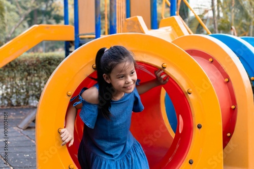 Young Asian elementary schoolgirl crawling through a colorful tunnel on a playground. weekend carefree holiday in park. bright playground equipment and focus expression capture childhood exploration