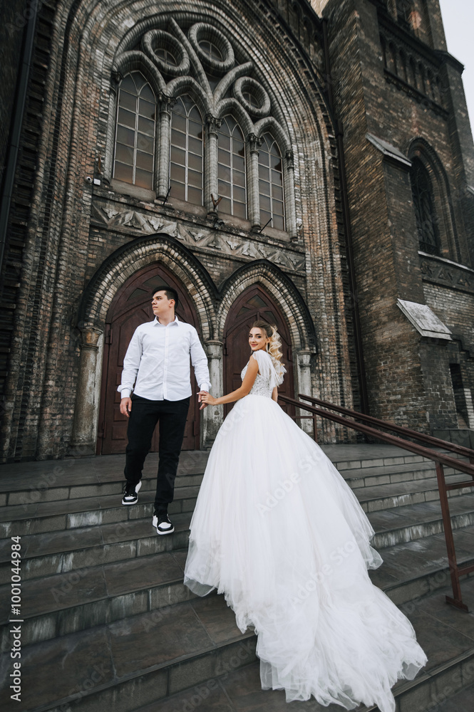 Stylish young groom and beautiful smiling bride curly blonde in white lace dress hugging, walking against the background of old brick church. Wedding photo, portrait of happy newlyweds. Stylish young groom and beautiful smiling bride curly blonde in white lace dress hugging, walking against the background of old brick church. Wedding photo, portrait of happy newlyweds.