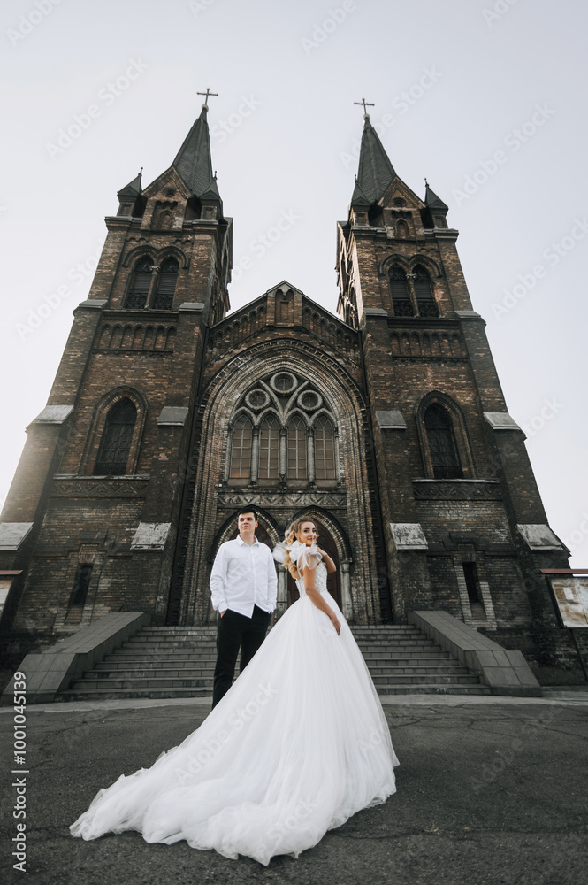 Obraz premium Stylish groom and beautiful blonde bride in white lace dress hugging standing against the background of old brick church. Wedding photo of happy newlyweds.