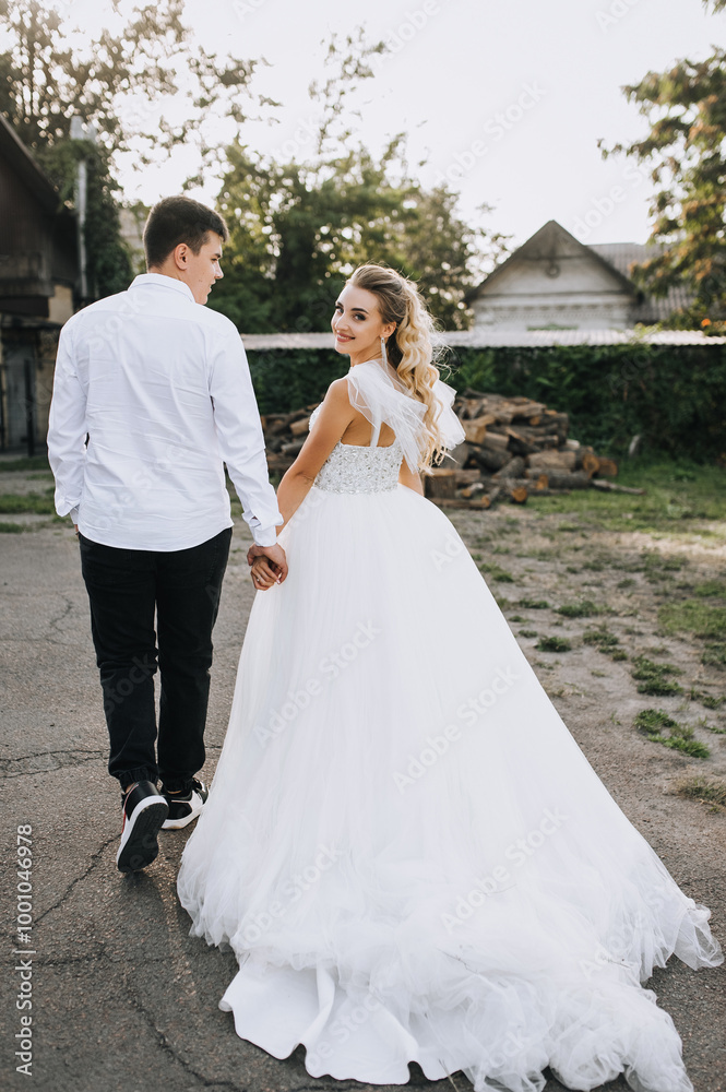 Naklejka premium Young groom and beautiful curly bride in white long dress walking holding hands outdoors against the background of logs in the village. Wedding photography, portrait of happy newlyweds.