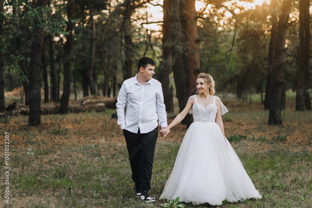 Stylish groom and beautiful smiling bride curly blonde in white long dress walking holding hands outdoors in forest in nature at sunset. Wedding photography, portrait of happy newlyweds.