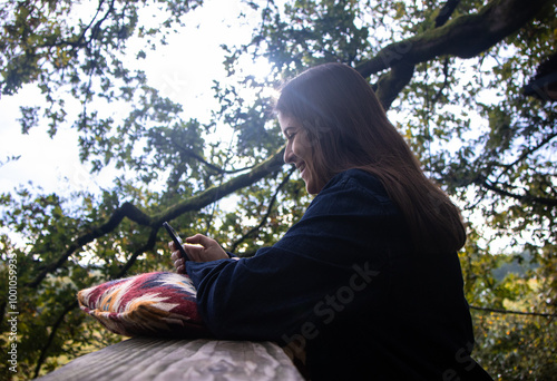 Profile view of a young brunette woman with Latin features smiling while looking at her cell phone screen, leaning on a nice cushion on the terrace of a house surrounded by green trees. #1001059933