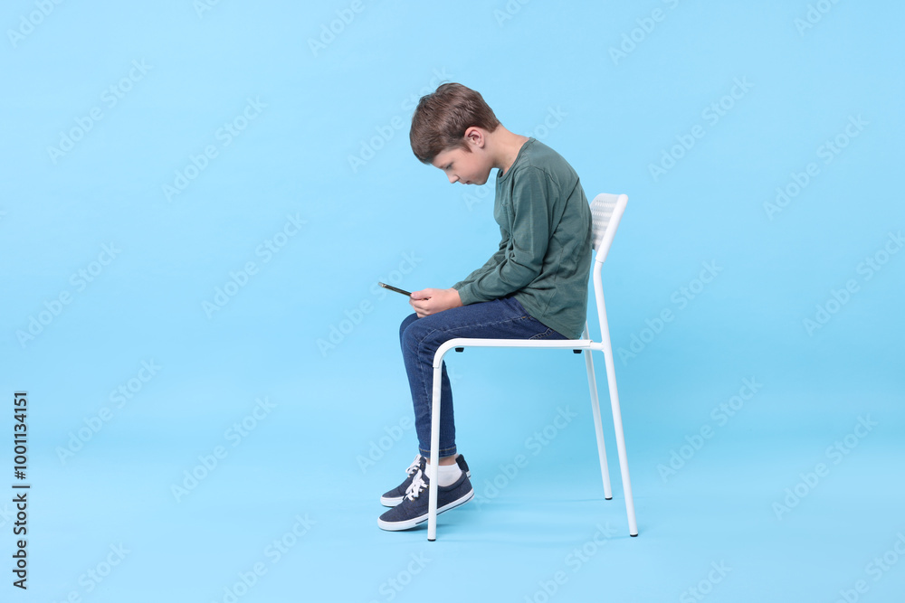 Boy with incorrect posture and phone sitting on chair against light blue background Boy with incorrect posture and phone sitting on chair against light blue background