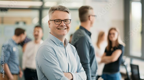 A smiling man in a white shirt stands confidently in a lively office setting, radiating positivity and approachability.