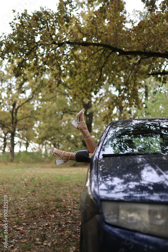 An artistic shot of a woman’s legs in stylish high-heeled sandals, playfully sticking out from the window of a parked car. The autumn trees create a serene, carefree vibe, blending nature and fashion