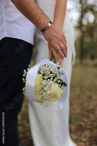 Close-up of a couple holding hands, with a delicate wedding bouquet of white roses and  tied with a satin ribbon. The subtle details of their rings and gentle touch symbolize love and commitment
