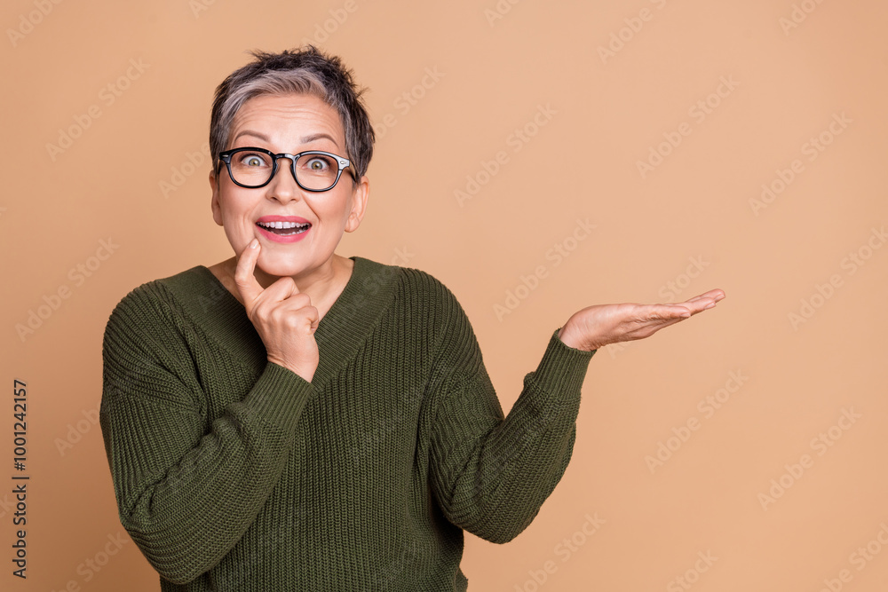 Photo of ecstatic woman with short hair dressed sweater in glasses hold product on palm empty space isolated on beige color background Photo of ecstatic woman with short hair dressed sweater in glasses hold product on palm empty space isolated on beige color background