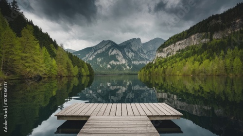 A serene photo of a lake nestled between lush green forests and towering mountains. The sky is overcast with dark clouds. In the foreground, a wooden dock extends over the water. The reflection of the