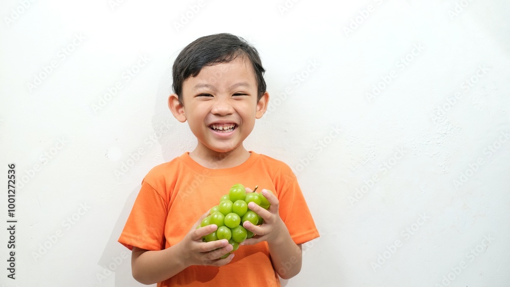 A cheerful boy in an orange shirt is holding a bunch of Shine Muscat grapes, his face expressing delight as he enjoys the sweet, juicy fruit. Perfect for promoting healthy eating and fresh produce. A cheerful boy in an orange shirt is holding a bunch of Shine Muscat grapes, his face expressing delight as he enjoys the sweet, juicy fruit. Perfect for promoting healthy eating and fresh produce.
