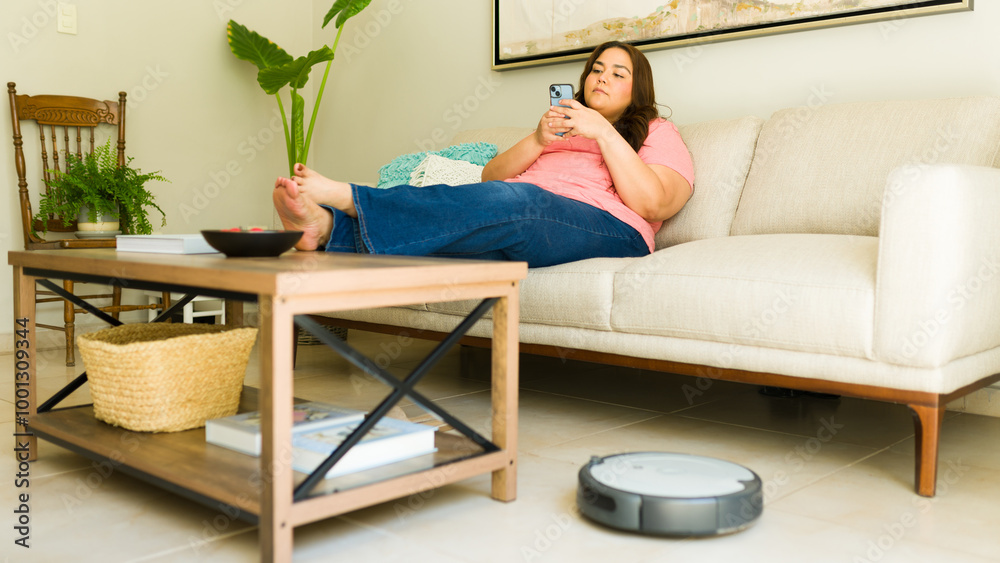 Plus size woman lounging on the sofa with her phone while a robotic vacuum cleans the living room Plus size woman lounging on the sofa with her phone while a robotic vacuum cleans the living room