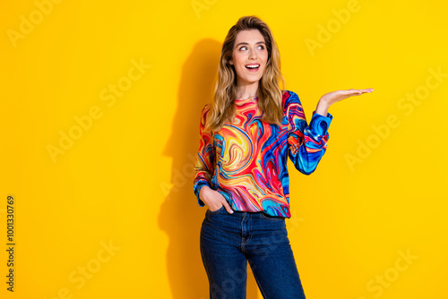 Photo of young shocked funky woman wearing colorful pullover and hold arm looking great deal isolated on yellow color background