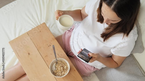 Hungry woman savoring healthy breakfast of organic oatmeal in bowl while resting in cozy bed checking e-mail engaging of browsing social media scrolling online photo