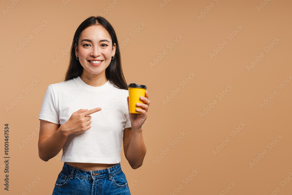 Young smiling Asian woman pointing at yellow take away coffee cup isolated on blue background Young smiling Asian woman pointing at yellow take away coffee cup isolated on blue background