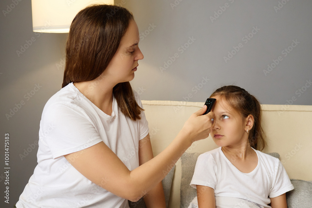 Concerned mother measuring her daughter's temperature with digital thermometer while little girl resting on couch feeling unwell from influenza and seeking care during her sickness at home Concerned mother measuring her daughter's temperature with digital thermometer while little girl resting on couch feeling unwell from influenza and seeking care during her sickness at home
