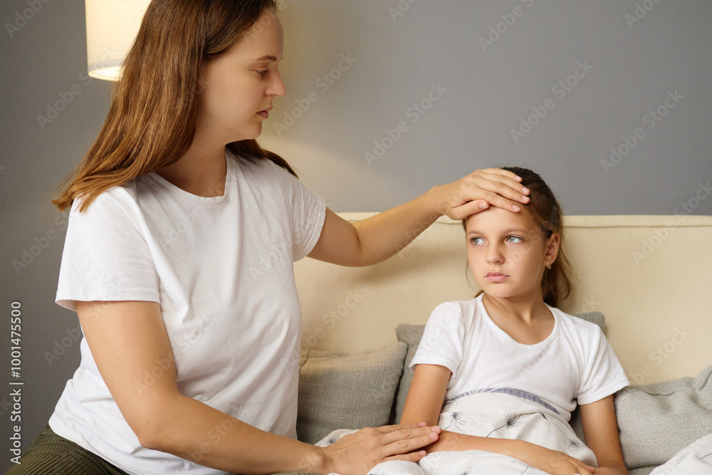 Fototapeta premium Sad upset mother checking her daughter's forehead for fever little girl sitting on the couch suffering from influenza symptoms while mother providing care for kid with grippe