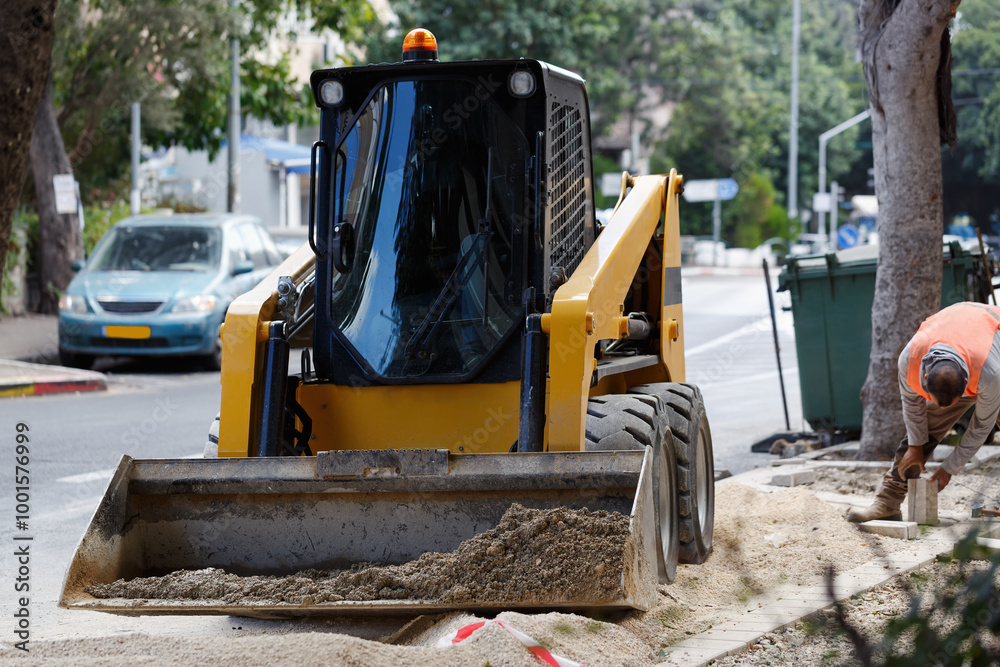 Yellow bulldozer on a street with its bucket lowered and filled with dirt. A worker in an orange vest is bending over, working on the sidewalk. Trees, parked cars are in the background. Construction Yellow bulldozer on a street with its bucket lowered and filled with dirt. A worker in an orange vest is bending over, working on the sidewalk. Trees, parked cars are in the background. Construction
