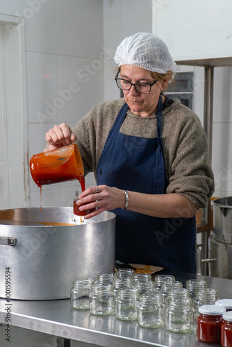 A worker in a jam factory pours freshly prepared Rosa Mosqueta (rosehip) jam into small glass jars. The production takes place in an artisanal setting, emphasizing the handcrafted quality. photo