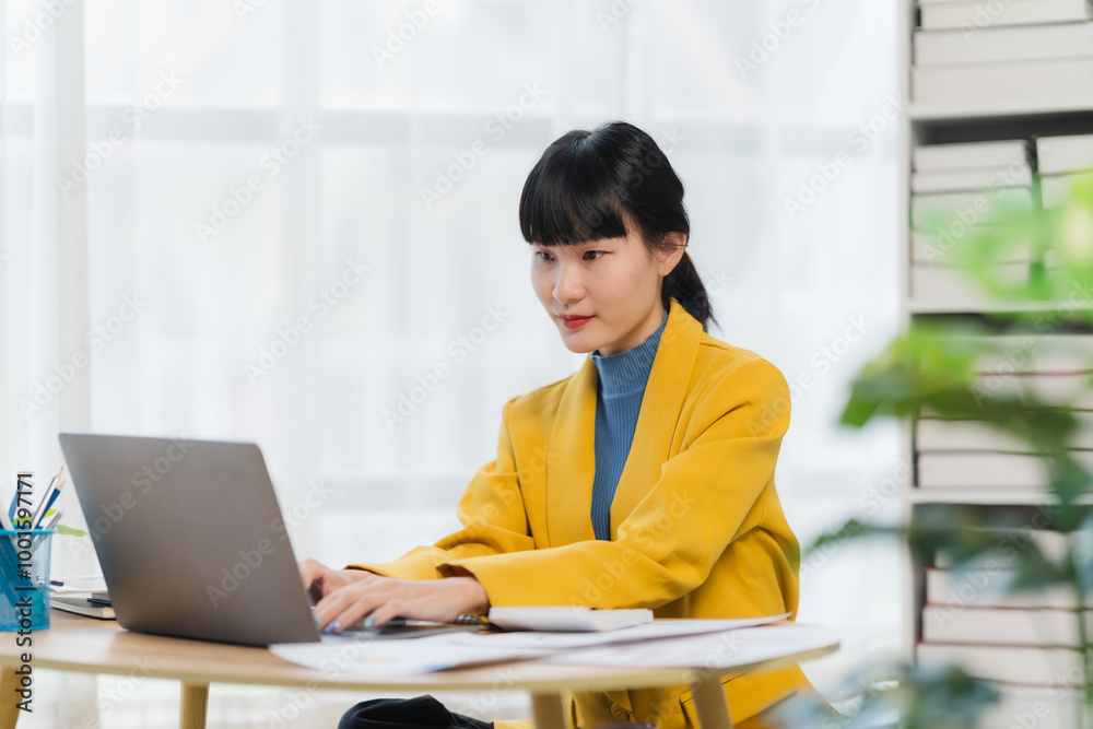 Focused and Ready: A young Asian businesswoman in a yellow blazer sits at her desk, intently focused on her laptop, radiating professionalism and determination. Focused and Ready: A young Asian businesswoman in a yellow blazer sits at her desk, intently focused on her laptop, radiating professionalism and determination.