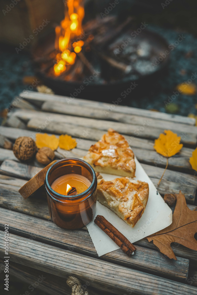Close-up of a lit candle in an amber glass jar, two slices of apple pie, and a cinnamon stick on a rustic wooden table. Autumn leaves and walnuts enhance the cozy atmosphere near a warm campfire Close-up of a lit candle in an amber glass jar, two slices of apple pie, and a cinnamon stick on a rustic wooden table. Autumn leaves and walnuts enhance the cozy atmosphere near a warm campfire