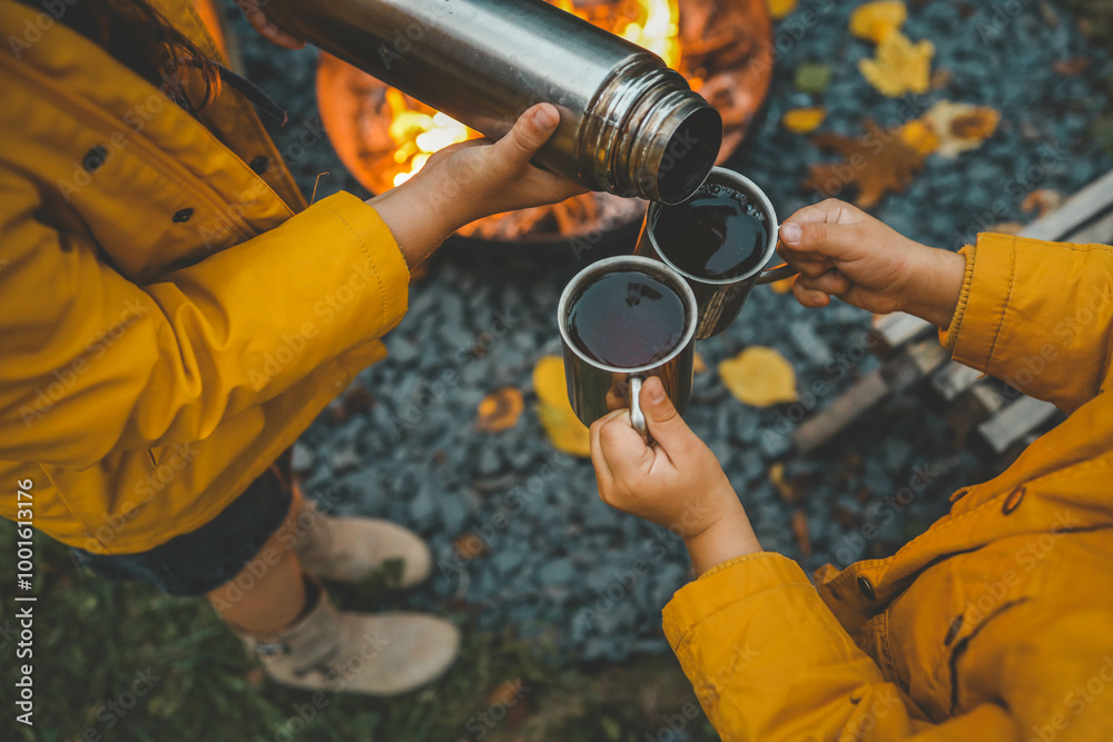 Naklejka premium Close-up people pouring hot drinks from thermos into metal mugs next to campfire. Cozy autumn scene with warm clothing, a woven basket, and the glow of the fire, creating a warm 