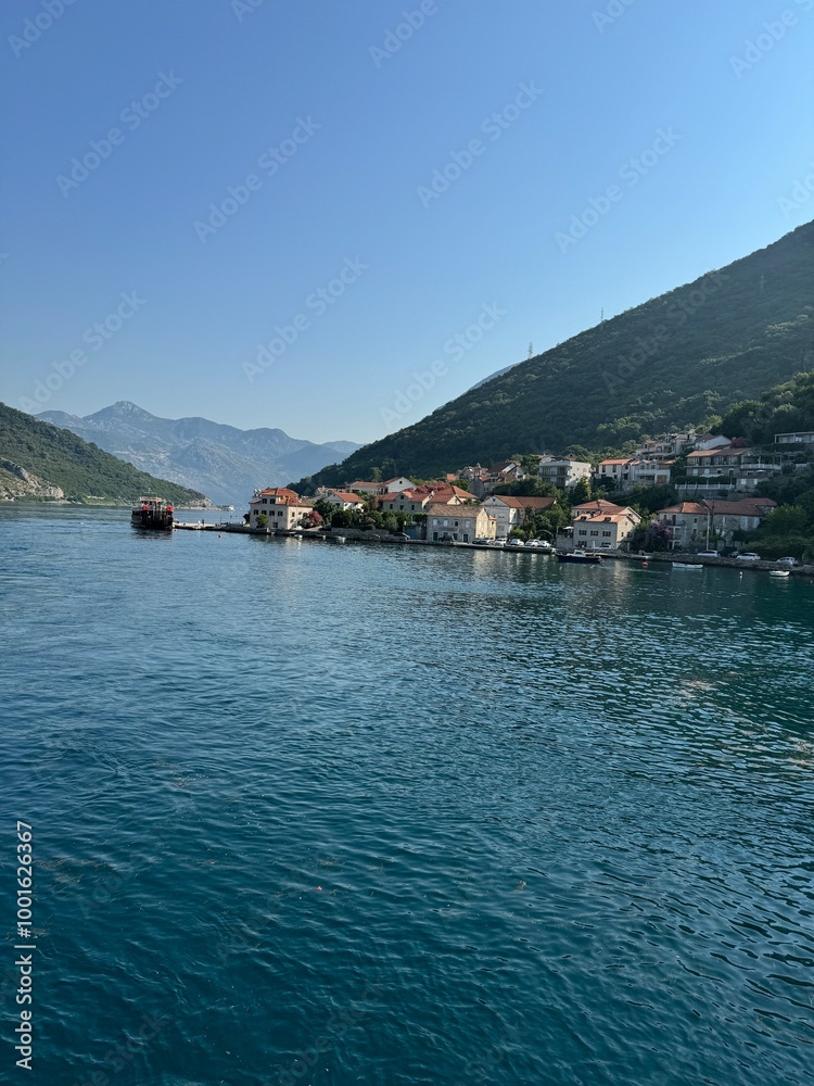 Scenic view of a coastal town nestled between mountains and the calm blue waters of a bay. The charming houses with red rooftops contrast against the lush green hills and clear sky