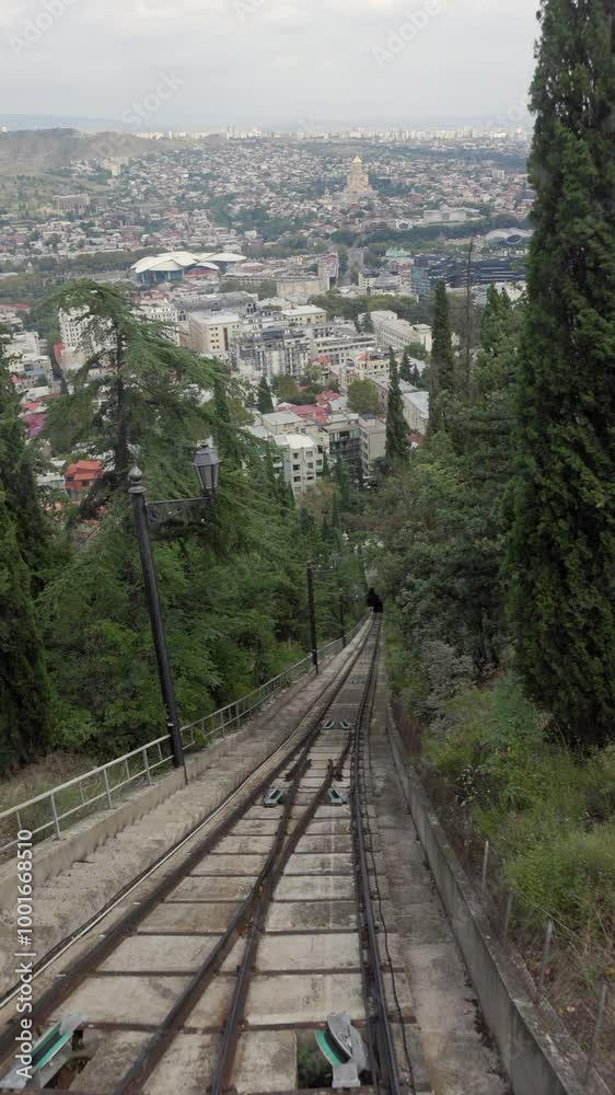 View from the cabin of the funicular car of the capital of Georgia Tbilisi, showcasing a vast array of architectural styles, a winding river, and prominent landmarks under a cloudy sky View from the cabin of the funicular car of the capital of Georgia Tbilisi, showcasing a vast array of architectural styles, a winding river, and prominent landmarks under a cloudy sky