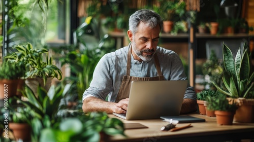 A middle-aged man in an apron focuses on his laptop while managing his online plant shop surrounded by various houseplants and gardening tools