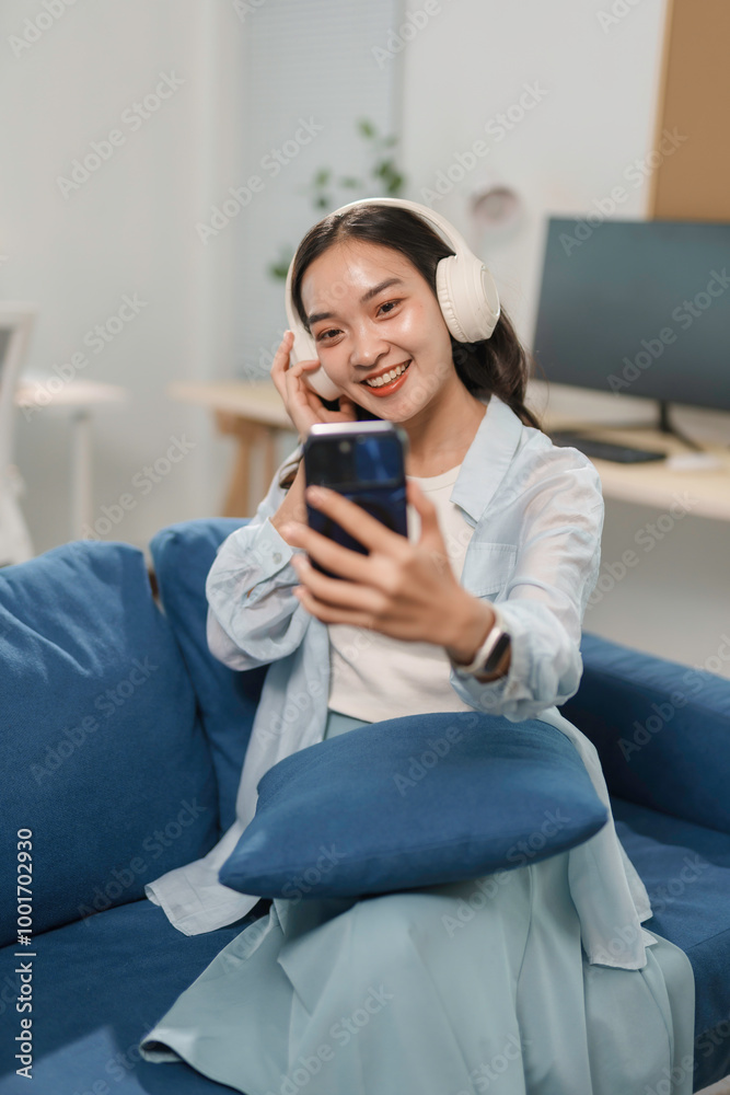 Young woman is sitting on a blue sofa at home, wearing headphones and using a smartphone Young woman is sitting on a blue sofa at home, wearing headphones and using a smartphone