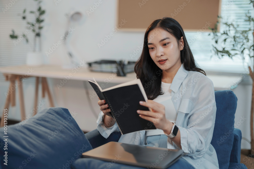 Young asian woman is sitting on a blue sofa in her home office, enjoying a moment of peace and quiet as she reads a book Young asian woman is sitting on a blue sofa in her home office, enjoying a moment of peace and quiet as she reads a book