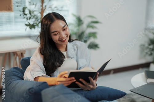 Smiling young woman sitting on a sofa reading a book, enjoying a relaxing leisure activity at home